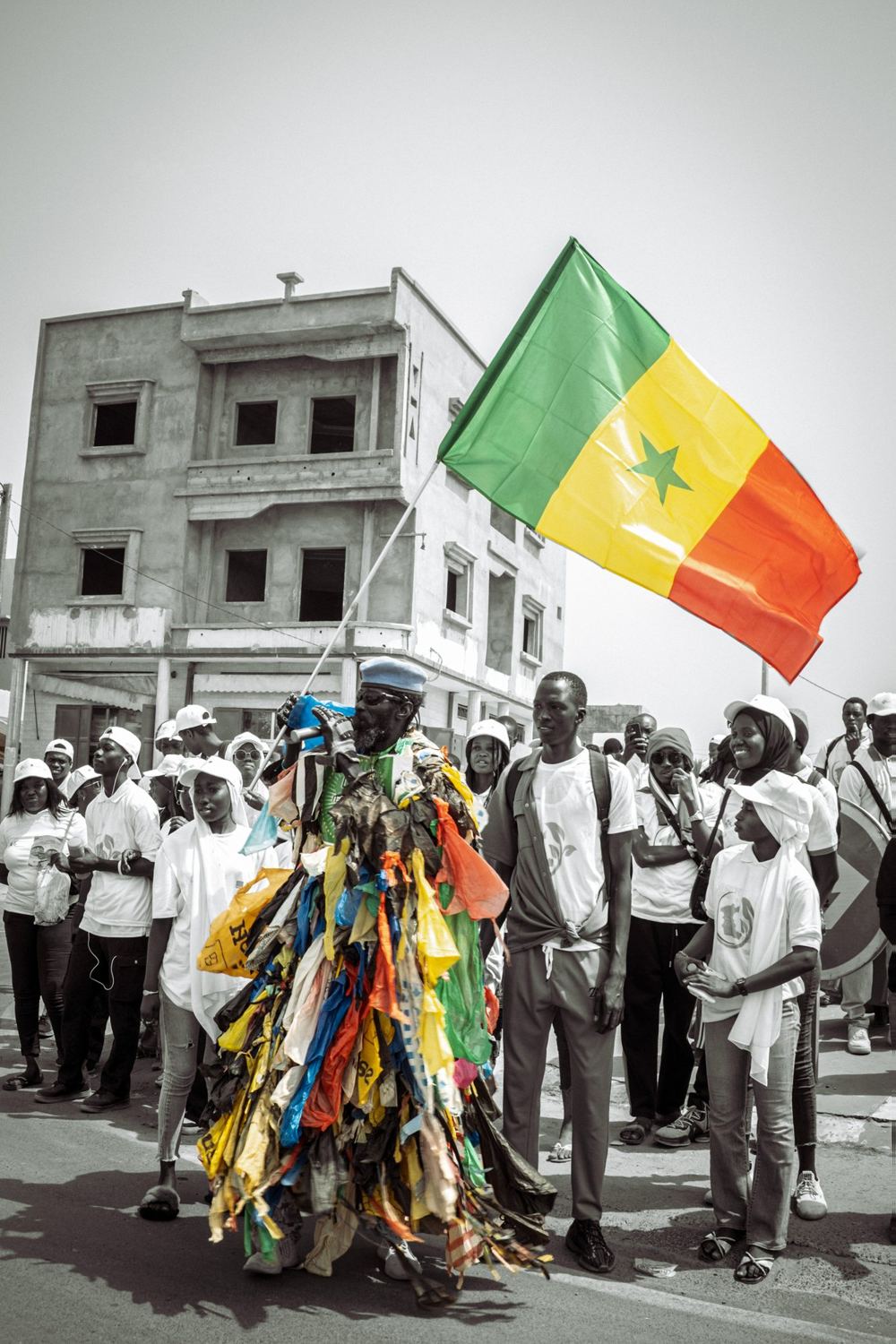 L’Homme Plastique with a large Senegalese flag in front of a crowd, the flag in colour, the rest in grey